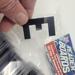 Puede incluir: Una pegatina negra con la letra "E" sobre un soporte transparente, sostenida por una mano. La pegatina es rectangular. Se ve una pila de pegatinas similares. Un kit de bricolaje Reader Boards est&aacute; en el fondo.