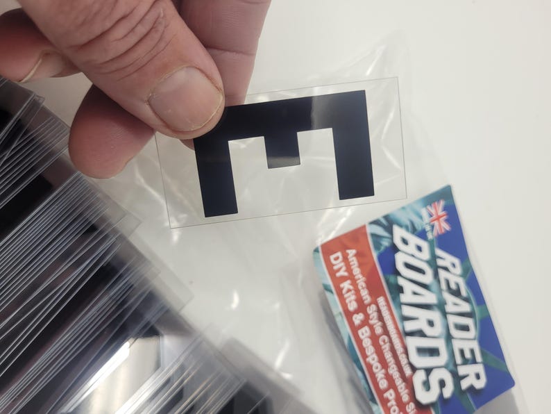 Puede incluir: Una pegatina negra con la letra "E" sobre un soporte transparente, sostenida por una mano. La pegatina es rectangular. Se ve una pila de pegatinas similares. Un kit de bricolaje Reader Boards est&aacute; en el fondo.