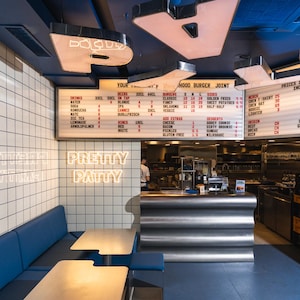May include: A restaurant interior featuring a blue bench seat, two light-coloured tables, and a metallic counter. A large illuminated menu board displays food and drink options, while a neon sign on the white tiled wall reads "PRETTY PATTY."