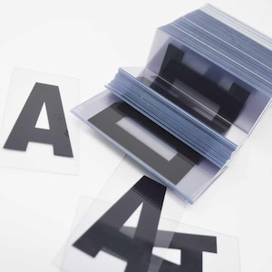 Puede incluir: Una pila de l&aacute;minas de pl&aacute;stico transparente con la letra "A" negra impresa. Varias letras "A" individuales est&aacute;n dispersas. Las letras est&aacute;n en una fuente negrita sin serifa.