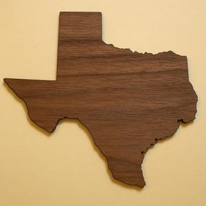 May include: A wooden cutout in the shape of the state of Texas. The wood has a dark brown color with visible grain patterns. The cutout is mounted on a light yellow background.