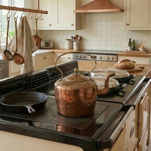 May include: A large, hammered copper tea kettle with a curved handle and spout sits on a black cast iron stove. Beside it, a black cast iron skillet rests on the stovetop. The kitchen features cream-coloured cabinets and a copper range hood.