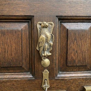 May include: A brass eagle door knocker mounted on a dark brown wooden door with raised panels. Below the eagle is a circular textured strike plate, a brass keyhole escutcheon with a metal key inserted, and a rectangular brass mail slot cover.