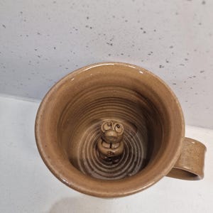 May include: A handmade brown ceramic mug with a sculpted creature inside. The mug has a handle and a textured interior with concentric circles. The creature has large eyes and a small mouth.