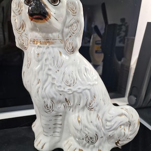 May include: A white ceramic dog figurine with gold accents. The dog is sitting upright with detailed fur and facial features. The figurine has a gold collar and decorative gold swirls. The dog's nose is black, and its eyes are brown.