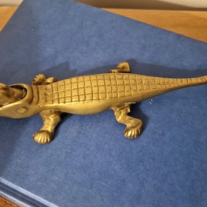May include: A gold-coloured alligator-shaped bottle opener. The opener has textured scales and is on a blue surface. The alligator has four legs and a long tail. The item is likely a decorative item.