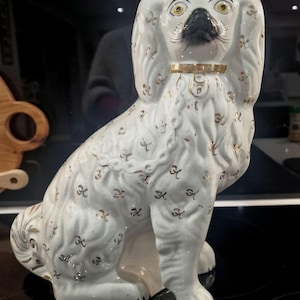 Pode incluir: Estatueta de cão em cerâmica branca com detalhes dourados. O cão está sentado com uma coleira dourada e um nariz preto. A estatueta tem detalhes dourados por todo o corpo.