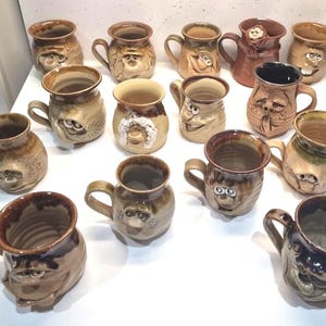 Op de afbeelding: Een verzameling handgemaakte keramische mokken met grillige, expressieve gezichten. De mokken zijn in verschillende bruin- en beigetinten, met unieke gezichtskenmerken en handvatten. Perfect voor een unieke keukenverzameling.