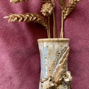 May include: A handcrafted ceramic vase with a light blue and brown glaze, featuring sculpted leaf and flower details. The vase contains dried wheat stalks, forming a rustic, natural arrangement.