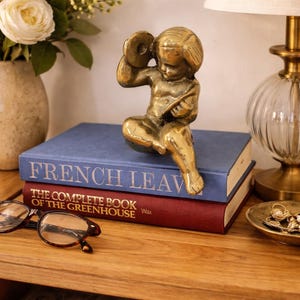 Könnte beinhalten: Eine Messing-Figur eines musizierenden Engels sitzt auf zwei Büchern. Das obere Buch ist blau mit dem Aufdruck "FRENCH LEAV". Das untere Buch ist rot mit dem Aufdruck "THE COMPLETE BOOK OF THE GREENHOUSE".