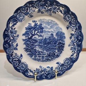 May include: A white and blue decorative plate with a scalloped edge. The plate features a blue floral border and a central landscape scene in blue. The landscape depicts trees, buildings, and figures. The plate is displayed on a gold-colored stand.