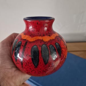 Pode incluir: Um pequeno vaso de cerâmica redondo com acabamento brilhante. A cor base é um vermelho vibrante com manchas pretas. Apresenta uma faixa horizontal laranja perto do pescoço e uma série de padrões verticais escuros em forma de gota.