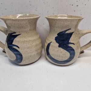 Op de afbeelding: Twee beige keramische mokken met een gespikkelde afwerking en gebogen handvatten. Elke mok heeft een donkerblauw abstract ontwerp op de voorkant. De mokken hebben een brede opening en een licht uitlopende rand. Deze mokken zijn geschikt voor koffie of thee.
