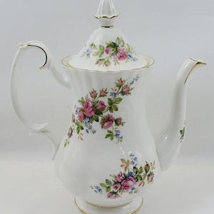 Könnte beinhalten: Weiße Porzellan-Kaffeekanne mit Deckel, Henkel und Ausguss. Die Kanne ist mit rosa Rosen, blauen Blumen und grünen Blättern verziert. Goldfarbene Verzierungen akzentuieren den Henkel, den Ausguss und den Sockel. Ein klassisches Design zum Servieren von Kaffee oder Tee.