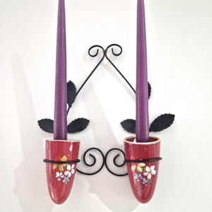 May include: A wall-mounted candle holder featuring two purple taper candles. The red ceramic cups are adorned with floral patterns and held by a black metal frame with leaf and scroll details. The candles are unlit.
