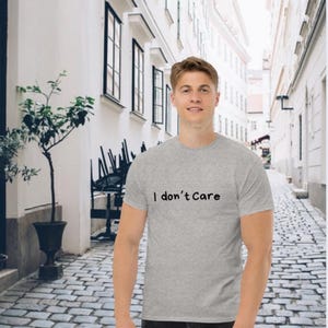Könnte beinhalten: Ein hellgraues T-Shirt mit dem schwarzen Aufdruck "I don't Care". Das kurzärmelige Shirt scheint aus einem weichen Material zu sein. Die Person steht vor einer Kopfsteinpflasterstraße.