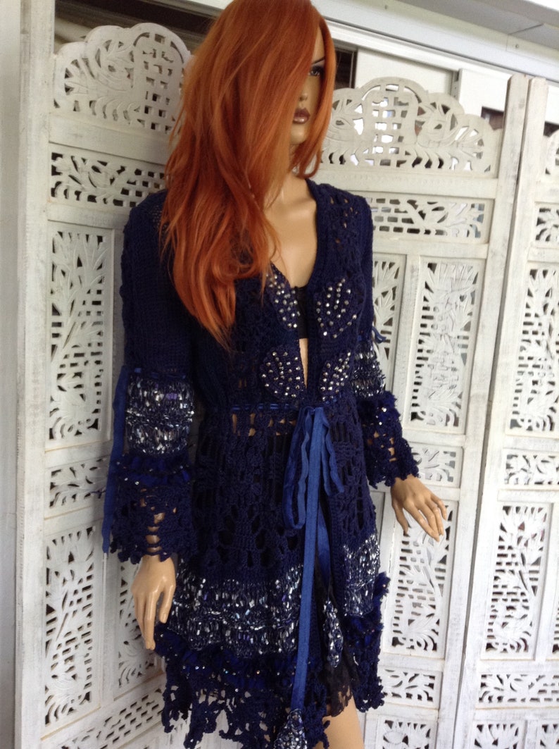 May include: A navy blue crochet cardigan with a beaded design and a blue ribbon tie. The cardigan has a long, flowing silhouette and a delicate lace trim.