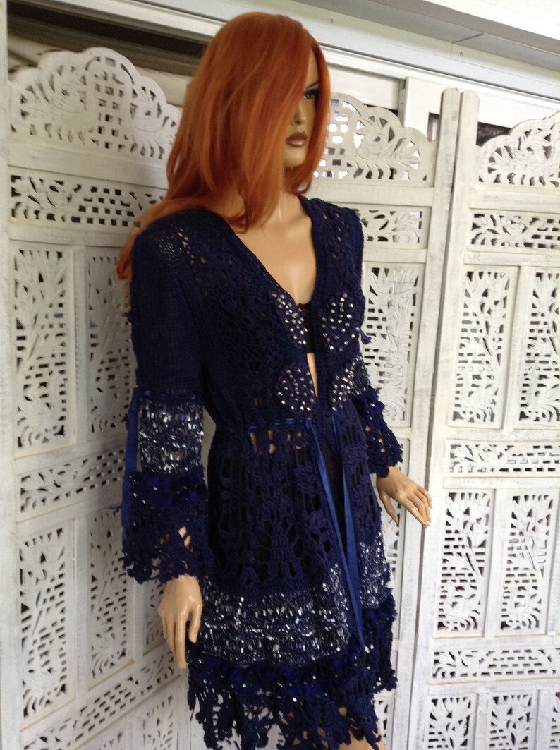 May include: A navy blue crochet cardigan with a long, open front and a lace trim. The cardigan is decorated with silver sequins and has a blue ribbon tie.