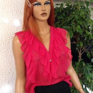 Puede incluir: Una blusa sin mangas, de color rosa brillante y semitransparente, con un escote en V profundo y volantes en cascada a lo largo de la tapeta frontal. La blusa se cierra con un botón visible y se combina con una falda negra texturizada de capas.