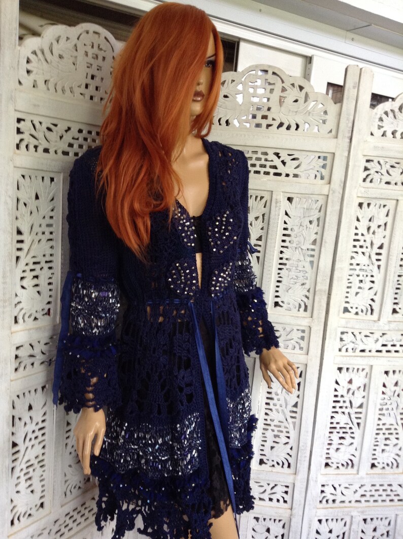 May include: A navy blue crocheted cardigan with a lace pattern and beaded embellishments. The cardigan has a long, flowing silhouette and a tie at the waist.