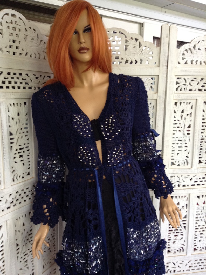 May include: A navy blue crocheted cardigan with a long, open front and long sleeves. The cardigan is embellished with silver sequins and rhinestones. It has a blue ribbon tie at the waist.