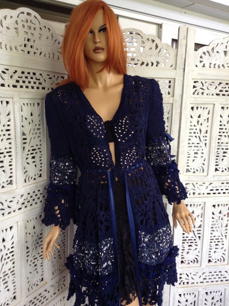 May include: A navy blue crocheted cardigan with a lace pattern and silver embellishments. The cardigan has a V-neckline and long sleeves with a ruffled trim.