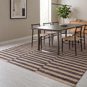 Peut inclure: Un tapis rectangulaire présentant un motif répétitif de rayures horizontales noires et beige naturel. Le tapis est tissé avec une finition texturée et recouvre une grande partie du sol sous une table de salle à manger en bois foncé et six chaises noires.