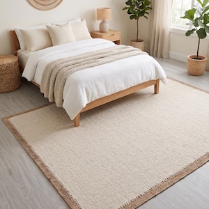 May include: A bedroom scene featuring a light-colored rug with a brown border, a wooden bed frame, and a white comforter. The room is decorated with neutral tones, including a woven mirror, a bedside table, and potted plants, creating a calming atmosphere.