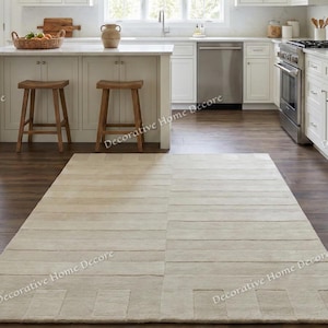May include: A rectangular, light beige rug with a geometric pattern is laid on a dark wood floor in a kitchen. The rug's design features horizontal and vertical lines, creating a textured appearance. The kitchen has white cabinets, stainless steel appliances, and wooden stools.
