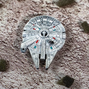 Puede incluir: Modelo detallado en plata del Halcón Milenario con detalles en rojo y azul. La réplica de la nave espacial presenta intrincados paneles y elementos de diseño, capturando el aspecto icónico de la nave espacial de Star Wars. El modelo está sobre un fondo texturizado y neutro.