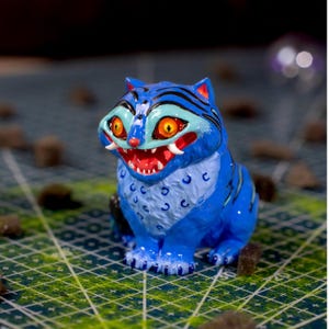 Puede incluir: Una figura de tigre de cerámica azul y blanca con una expresión feroz. El tigre tiene ojos naranjas, boca roja y colmillos blancos. Rayas negras adornan su cuerpo. La figura está sobre una superficie de cuadrícula verde y blanca.