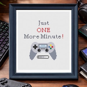 Puede incluir: Arte de punto de cruz enmarcado con temática de juegos. La obra presenta un mando de juego pixelado gris y el texto "Just ONE More Minute!" en rojo y gris. El marco es azul oscuro y el fondo es blanco.