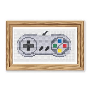 May include: Cross-stitch artwork of a classic video game controller in gray, black, and primary colors. The controller is framed in a light brown wooden frame, set against a white background. The design features a directional pad, buttons, and a start button.