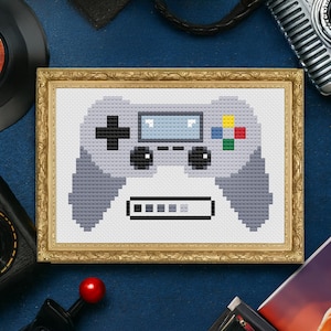 May include: Cross-stitch artwork of a gray video game controller in a gold frame. The controller features a directional pad, buttons, and a screen. The frame is rectangular with a decorative gold border. The background is a dark blue.