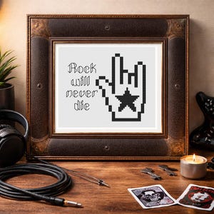 May include: Framed cross-stitch art with the text "Rock will never die" and a pixelated hand gesture. The frame is brown with a decorative edge. The artwork is on a white background. A black cable and headphones are in the foreground.