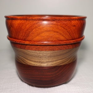 May include: A handcrafted wooden bowl with a layered design. The top section is a rich reddish-orange, followed by a lighter tan band, and a deep reddish-brown base. The bowl has a smooth, polished finish, showcasing the natural wood grain.