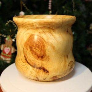 Pode incluir: Uma tigela de madeira artesanal com uma cor castanha clara e um padrão de grão de madeira único. A tigela tem uma forma arredondada com uma abertura larga e uma borda ligeiramente alargada. O grão da madeira apresenta um proeminente design de nó.