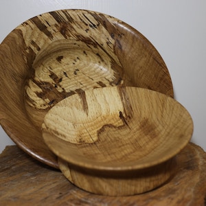 Puede incluir: Dos cuencos de madera de diferentes tamaños, uno dentro del otro. Los cuencos son de madera marrón claro con vetas marrones más oscuras. El cuenco más grande es más ancho y menos profundo, mientras que el más pequeño es más profundo.