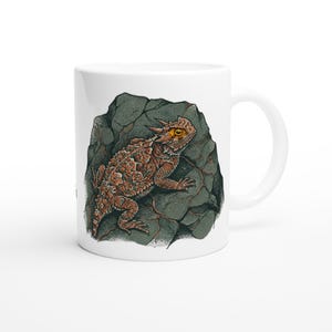 May include: White ceramic mug with a detailed illustration of a horned lizard on a grey rock. The lizard is depicted in shades of brown, orange, and yellow, with a textured appearance. The mug has a standard handle and a glossy finish.