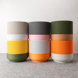 Könnte beinhalten: Eine Sammlung von sechs zylindrischen Pflanzgefäßen in verschiedenen Farben. Jedes Pflanzgefäß hat eine gerippte Textur auf dem oberen Teil und eine einfarbige Basis. Farben sind Beige, Schwarz, Olivgrün, Gelb, Grau und Orange.