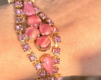 May include: A gold-toned bracelet with three rows of pink gemstones. The bracelet features a variety of pink stones, including round, teardrop, and square shapes. The bracelet is worn on a wrist, with a blurred background.