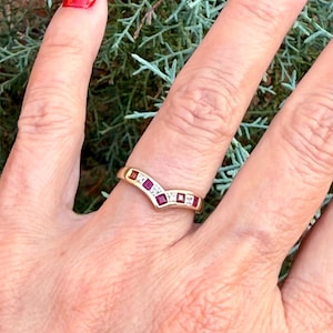 May include: A gold V-shaped chevron ring featuring a series of alternating square-cut red rubies and small clear diamonds set along the band. The ring has a polished metal finish and follows a pointed, angular design that sits on the finger.