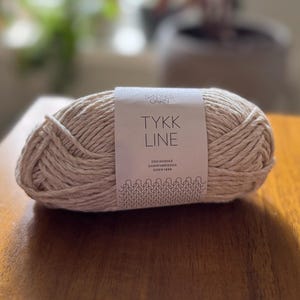 May include: A ball of beige yarn with a label that reads "TYKK LINE". The yarn is tightly wound and appears soft. The label also includes the text "DEN NORSKE GARNFABRIKKEN SIDEN 1888". The yarn is on a wooden surface.