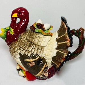 May include: A ceramic teapot shaped like a turkey with a glossy finish. The turkey has a deep red head, a yellow beak, and brown and cream feathers. The lid features sculpted acorns and leaves, while the handle resembles a textured brown tree branch.
