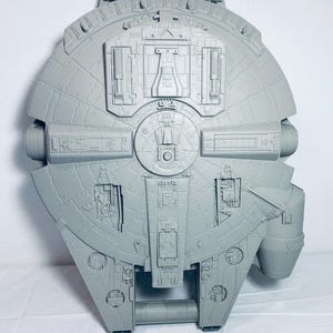 May include: A light gray plastic toy spaceship with a circular main body and a protruding cockpit on the right side. The surface features intricate molded mechanical details, including rectangular panels, vents, and geometric patterns across the hull.