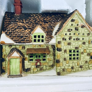 May include: A ceramic miniature cottage featuring a light green stone facade, a dark brown textured roof with a chimney, and a light green front door. The structure includes a small window with a green wreath and a red bow, set against white foam.