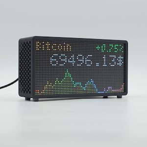 Peut inclure: Un &eacute;cran d'affichage num&eacute;rique rectangulaire noir avec un motif lat&eacute;ral en nid d'abeille. L'&eacute;cran affiche le texte Bitcoin, la valeur 69496.13$, et un indicateur vert plus 0.75% au-dessus d'un graphique lin&eacute;aire pix&eacute;lis&eacute; color&eacute;. Un c&acirc;ble noir d&eacute;passe.