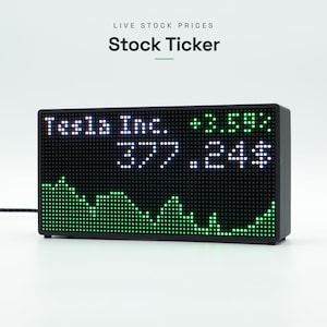 以下が含まれることがあります： ドットマトリクスLEDディスプレイを備えた長方形の黒いデジタル株価ティッカーデバイス。画面には「Tesla Inc.」、+3.59%の増加率、377.24ドルの価格、および市場トレンドを示す緑色の折れ線グラフが表示されています。