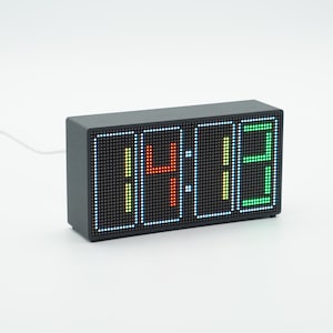 Puede incluir: Un reloj digital rectangular con carcasa gris oscuro y pantalla de matriz de puntos. La pantalla muestra la hora 14:13 con luces LED multicolores, con el primer dígito en blanco, el segundo en rojo, el tercero en amarillo y el cuarto en verde.