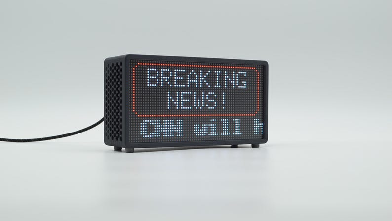 Peut inclure: Un appareil d'affichage num&eacute;rique LED noir rectangulaire avec un panneau lat&eacute;ral &agrave; motif en nid d'abeille. L'&eacute;cran affiche le texte BREAKING NEWS! en lettres blanches dans un cadre rectangulaire rouge, avec le texte partiel CHN will h en dessous.
